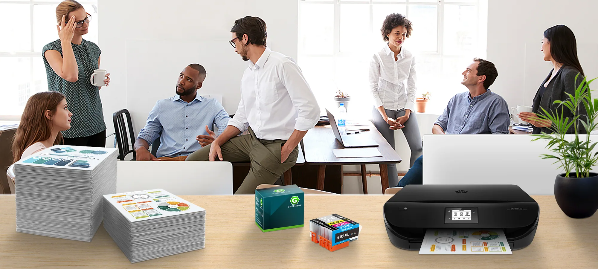 An office meeting behind a desk with stacks of printed pages, a green GREENBOX carton, ink cartridges and an inkjet printer