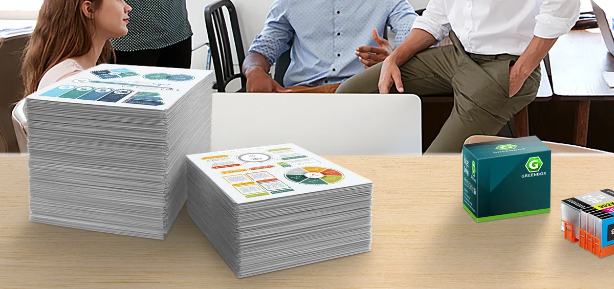Two stacks of printed color pages on a desk beside a green GREENBOX carton and ink cartridges